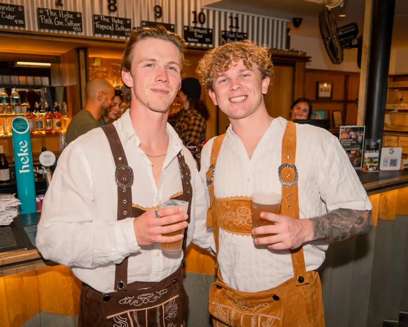 Two Oktoberfest guests in lederhosen holding drinks at the bar