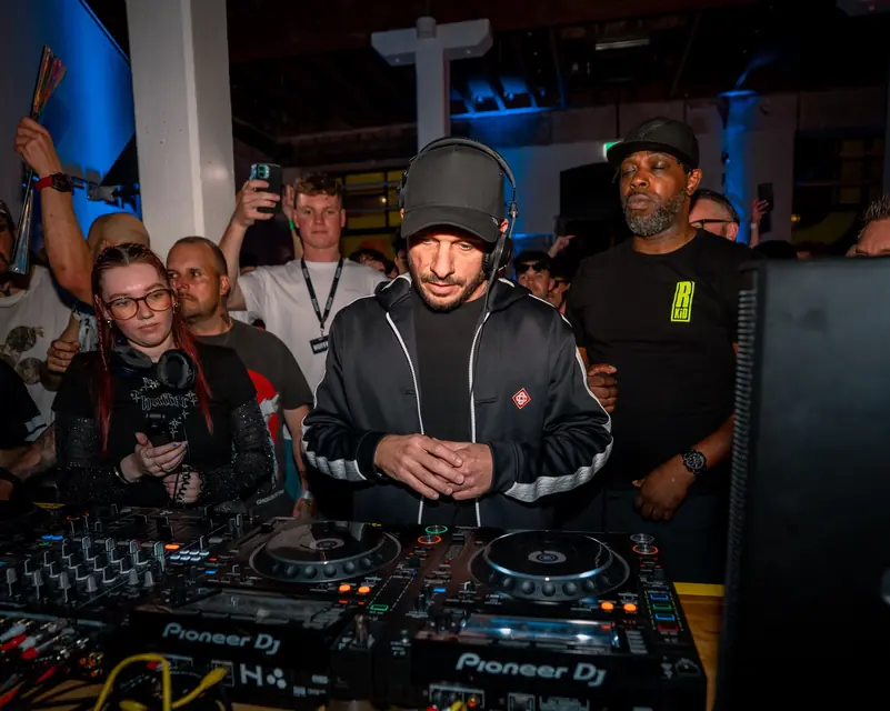DJ at crowded decks surrounded by an audience and other performers